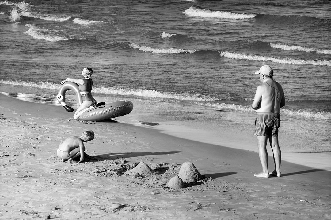 Bathers at the water’s edge beside sand figures and an inflatable ring.